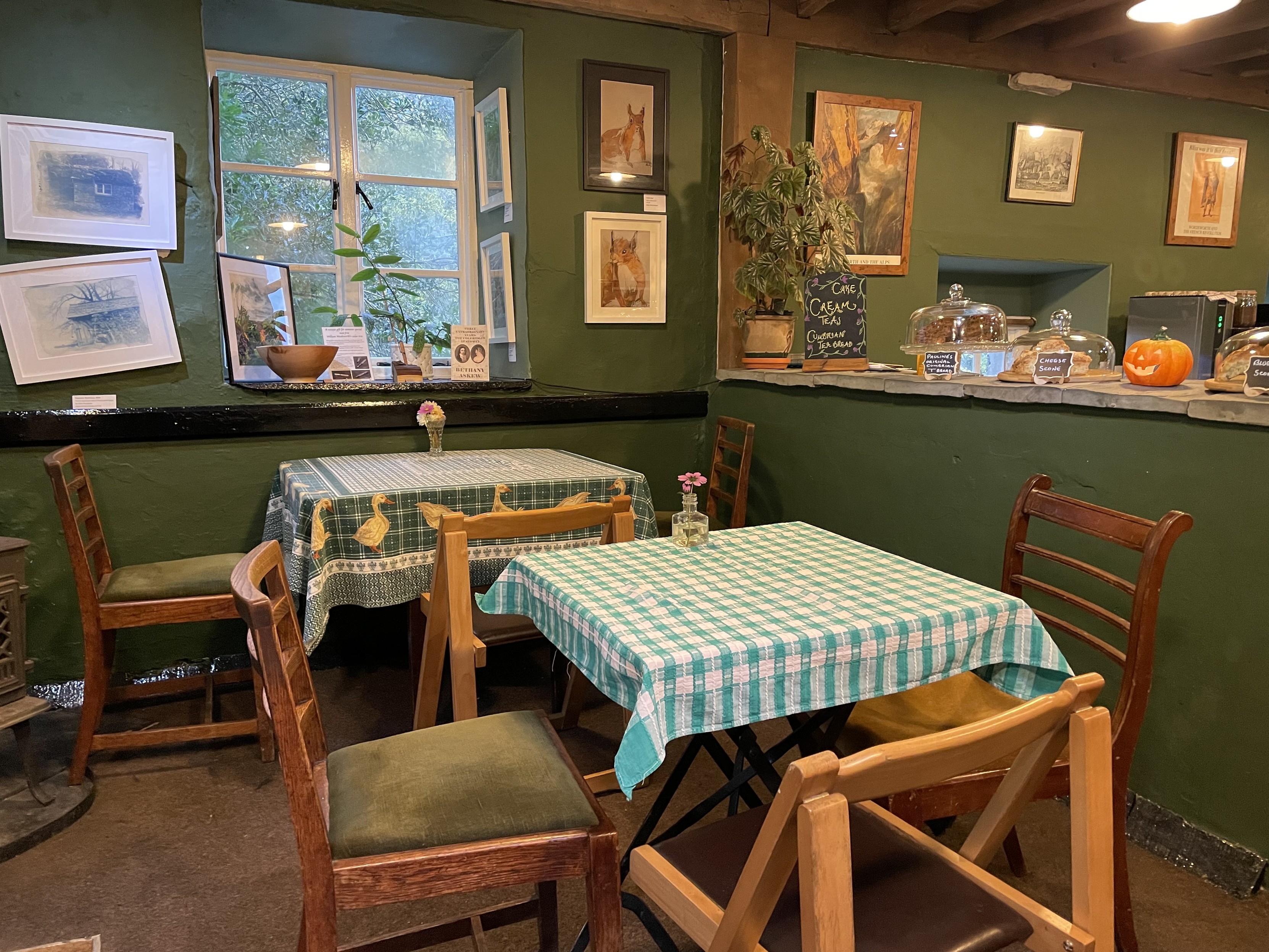 Two empty tables with three chairs each, covered by a green and white checkered table cloth, in a cafe with green walls with many paintings on them, next to a counter with various cakes on it under glass covers and a little pumpkin. 