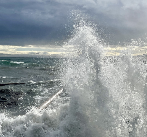 Waves hitting a seawall and sending spray many feet upwards, large logs being tossed in the water , breaking clouds with sunshine in the background backlighting the spray