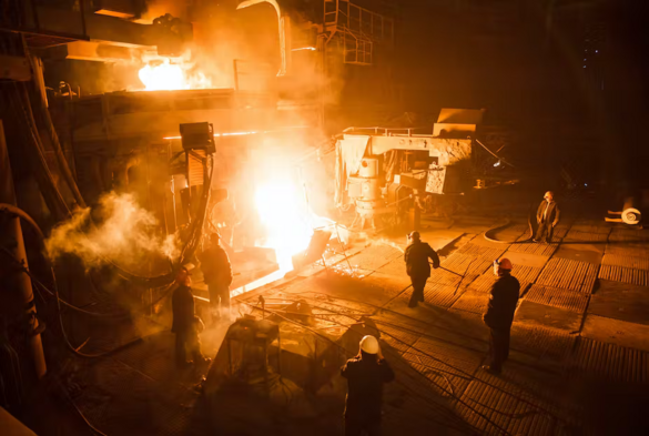 A group of workers operating an electric arc furnace inside a steelworks. The furnace is glowing very brightly.