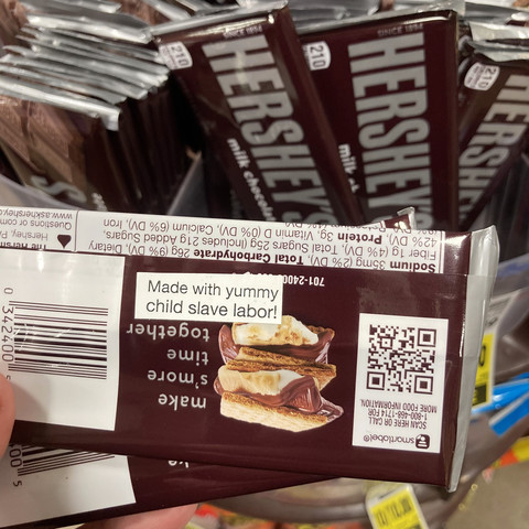 the back of a Hershey chocolate bar is held up against a backdrop of other Hershey bars on sale at a grocery store. there is a white sticker on the bar being held up that reads, "Made with yummy child slave labor!"