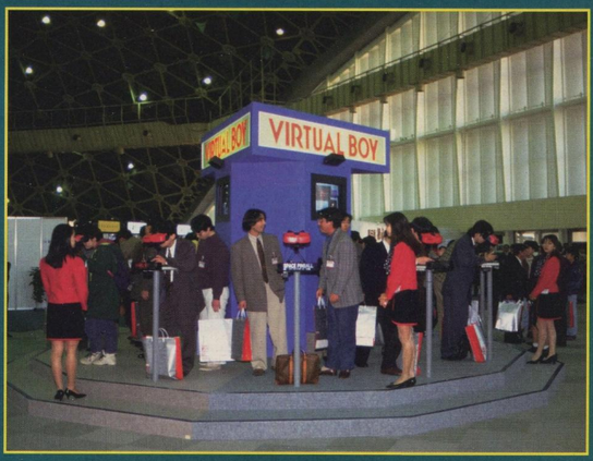 a circular set of steps with a blue-purple stand in the middle, with a 'virtual boy' sign up top on each of the four sides. virtual boys are set-up on poles at equal intevals around the inside of the top step and screens are displays on the sides of the stand. many people are milling around the area.