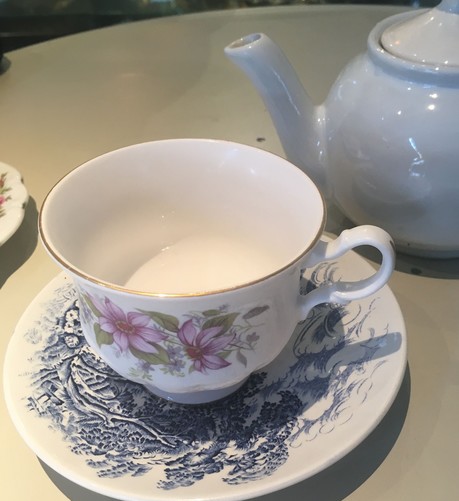 A flowery tea cup with a gold rim on a blue and white china saucer. Both are on a white table with a white teapot in the background. The tea cup does not contain any tea.