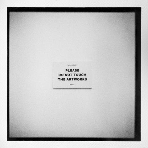 Photo of a sign mounted on a wall in a gallery saying "Please do not touch the artworks".