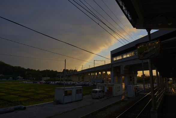 Later afternoon light silhouettes a raised Shinkansen train line. A bank of cloud covers most of the sky, its leading edge converging with the diagonal of the tail line in the distance. Long bands of light cast between the uprights of the raised train line illuminate cultivated farm beds to the image’s left, possibly growing rice.