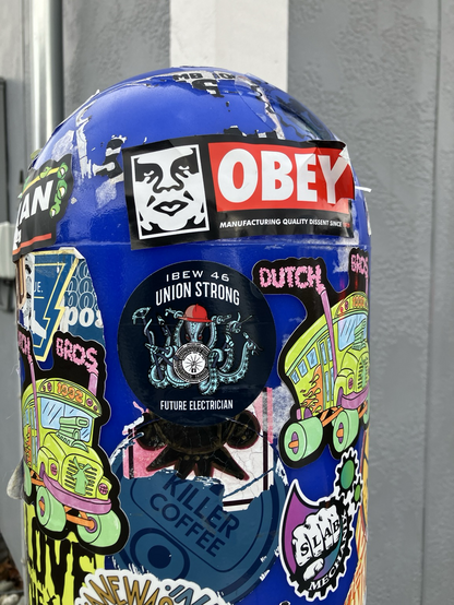 Bollard with many stickers: at center, an IBEW 46 “union strong” circle with an octopus in a hard hat and holding  a tool in each tentacle 