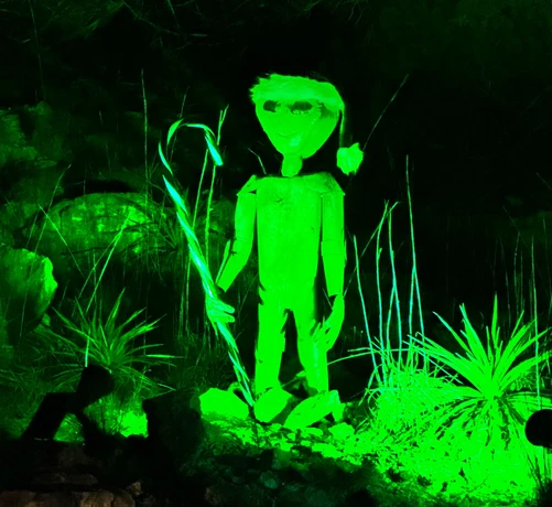 A metal yard art alien, with a green light spotlighting it. The alien is holding a candy cane and wearing a Santa hat.