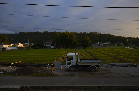 Cultivated fields in the midground, with horizontal bands of light and shadow being cast by a structure from out of frame. In the foreground is a small tip-truck on an unpaved road, where a drainage trench i being excavated next to the field. In the background, are forested hills.