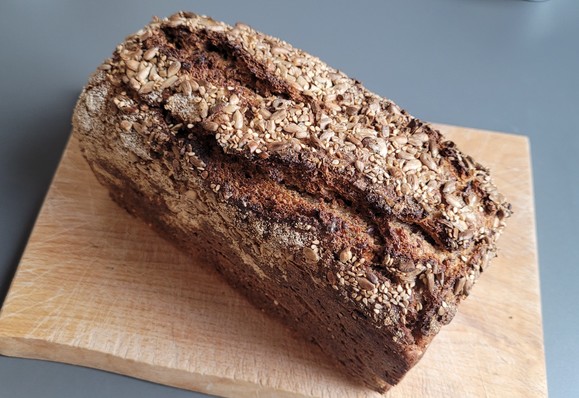 Farbfoto: Auf einem Schneidebrett liegt ein kastenförmiger Laib Brot, oben mit Sesam und Sonnenblumenkernen bestreut und kräftig aufgerissen. 