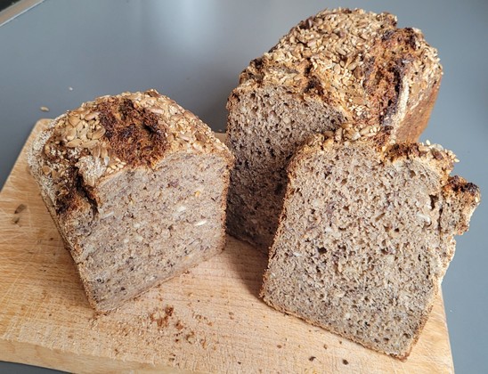 Farbfoto: Derselbe Brotlaib, teilweise aufgeschnitten, so dass man die fein bis mittelporige Krume mit Saaten (Leinsaat, Sonnenblumenkerne) sehen kann. Die Haferflocken sind nicht mehr erkennbar. 