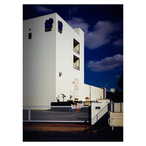 This image features a modern building with the logo of NGK, known for their spark plugs, affixed to its clean, white facade. The structure is highlighted by sunlight against a contrasting backdrop of dark blue skies, evoking the warmth and precision of technology at work.