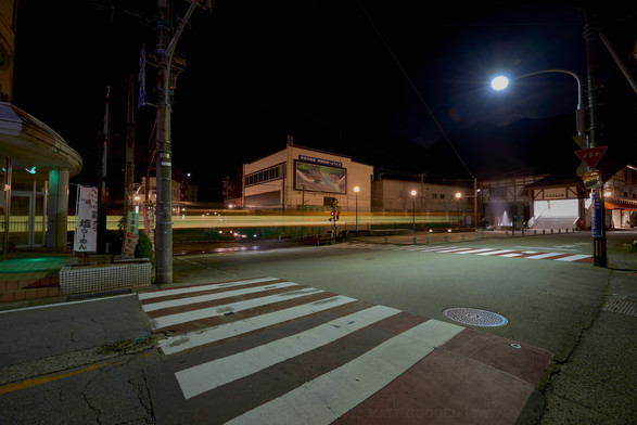 An intersection at night with a streetlamp showing in the sky above background mountains, looking lie the moon. Streaks of light are moving through the scene as a result of a long exposure of the local train passing.