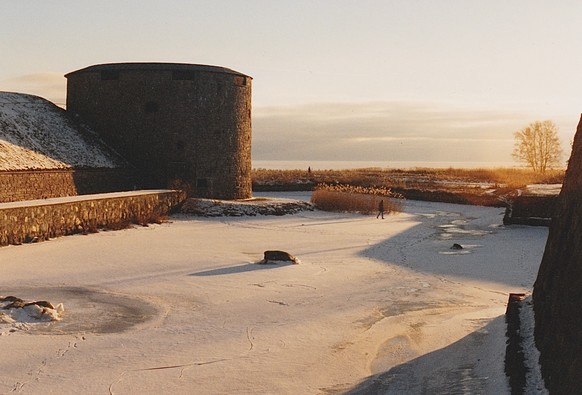 A winter view at Kalmar Castle in south eastern Sweden. This is the frozen moat. There is a stubby cylindrical tower, you can see the sea in the far background, and in the mid-ground you can see someone walking on the ice. It is late afternoon and the light is golden.