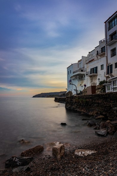 Se puede ver una orilla de mar en primer término, a la derecha casas del pueblo de Cadaqués (Girona) y al fondo un cielo de atardecer.

You can see a shore in the foreground, a House of the village of Cadaqués (Girona) in the right and a sunset Sky in the background.