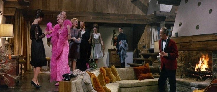David Niven in a red tux seated by a conversation pit and a host of high class entourage arriving in luxury resort in the 1960s. Screenshot from "The Pink Panther".