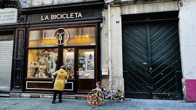 Woman in yellow down coat gazing at windows of a shop called "La Bicicleta", with a bicycle in front, its wheels decorated with flowers.
