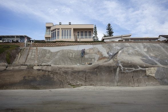 Image of a seawall being repaired by one worker on a ladder, sand in the foreground. 