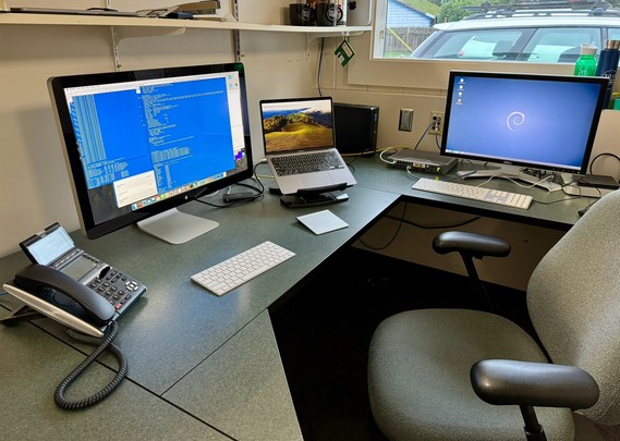 Office workspace with multiple computer monitors, a laptop on a stand, a keyboard, a mouse, a desk phone, and an office chair.