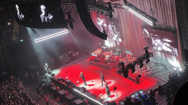Madness performing on stage at the O2 Arena. The stage is lit in red and there are big screens showing Suggs singing and holding out his left hand, Mike Bardon on keyboards, and Dan Woodgate on drums - all in black and white. 