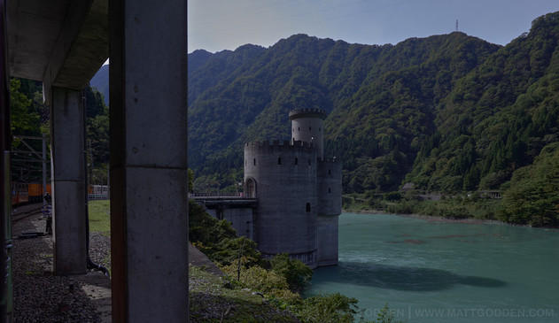 A… I don’t know what to call it. It’s a small European castle in the river, joined to the much higher bank by a bridge. I assume it’s for part of the hydroelectric infrastructure, but it’s a castl, in bright turquoise water, the far side of the gorge is almost to the top of the frame. On the far left, is the concrete structure of the tunnel the tourist train is travelling through periodically along the line.