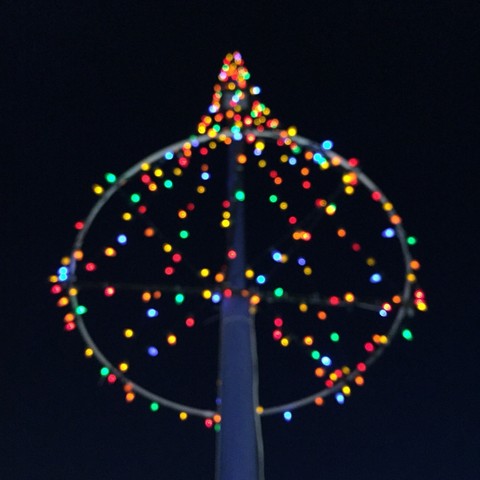 Square photo looking up at multicoloured lights outside in the dark. Lights hanging from a pole drape down around a circle base, making it look like a tree against a black background. The lights furthest away have a bokeh effect.
