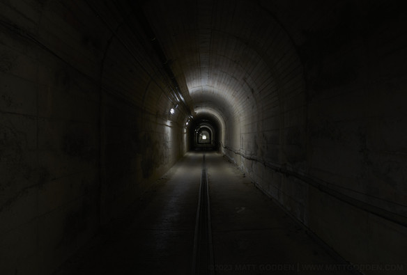 A concrete tunnel, with a tiny patch of light at the end. There are alternating bands of light and dark running around the tunnel, marking the location of each wall-mounted fluorescent tube lighting fixture. There is a central drainage grille running down the centre of the tunnel’s floor.