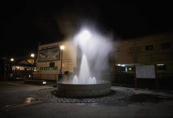 A night image of an approximately three meter circular fountain with a tall vertical central jet, surrounded by several smaller vertical jets. Above it, a cloud of heated steam hangs, like a shroud. The fountain is surrounded by cobblestones, and the background is lit with globular street lights.