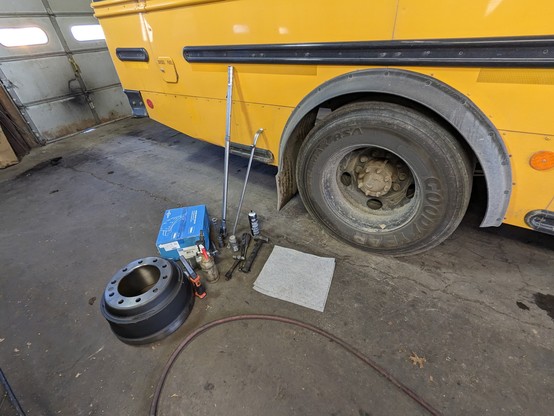 The rear wheel of a school bus. On the ground beside it is a new brake drum, a blue box with new brake shoes in it and various tools required for the job.