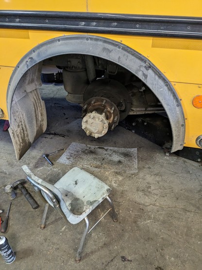 The rear wheel well of a school bus. Both wheels, the drum and brake shoes have all been removed, exposing the hub assembly. A small plastic school chair is sitting on the floor in front of it.