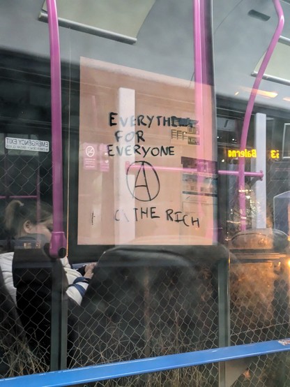 Through a bus window graffiti can be seen reading 'everything for every, eat the rich'