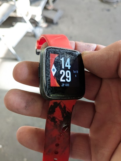 A Pinetime smart watch. I'm tilting it into the sunlight to highlight scratches all over the screen. It has a red band that is currently smeared with a combination of grease, brake dust and antiseize Liberian.