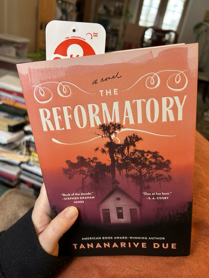 Me holding Tananarive Due’s new novel, The Reformatory in my hand. It has a squid bookmark over halfway through. In the background, piles of books that will one day be shelved more neatly, a year’s reading and projects.
