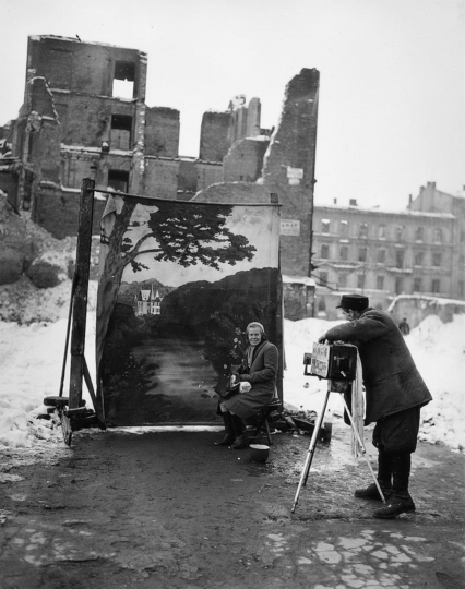describo la foto, ciudad destruida por la guerra, Polonia 1946, podría ser cualquier otra ciudad en la actualidad, una mujer posa sentada frente a un telón decorado que muestra un romántico paisaje con un sendero y un  palacio señorial al fondo, detrás del fotógrafo y el set improvisado tenemos a la ciudad destruida. Autor.   Michael Nash 1946