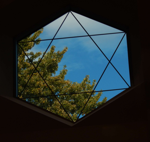 Photo: A hexagonal window with a six-pointed Star of David outlined in it. Outside the window is brilliant blue sky. Running from near the upper left side of the window to the lower right side are the branches of a tree. The branches are covered primarily by green leaves, although some light touches of autumn color — mostly orange — are beginning to show.