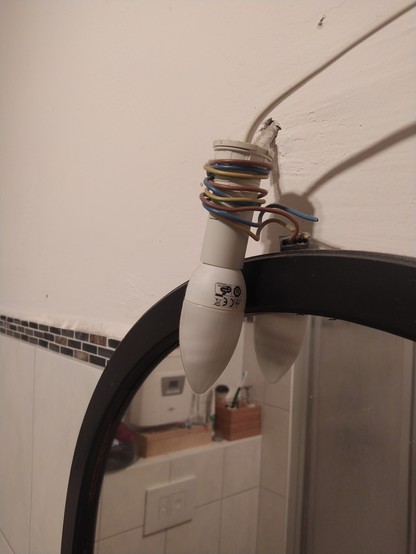 Eine nackte Lampe hängt mit Kabeln aus der Wand befestigt über einem Badezimmerspiegel