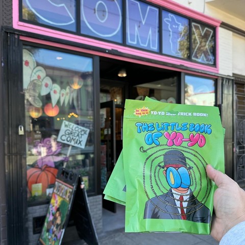 A photo of Silver Sprockets Comic Shop as seen from outside. In the foreground a hand is holding multiple copies of The Little Book of Yo-Yo. The Little Book of Yo-Yo is in a green plastic bag and it includes a comic book and a yo-yo inside each bag. 