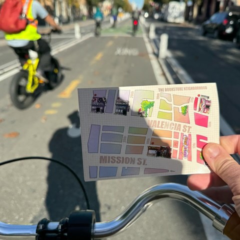 A photo I took while riding my bike down Valencia Street. I'm on my bike and holding out a postcard that says "The Bookstore Neighborhood.". The postcard shows a map of local bookstores on Valencia and Mission Street. There are several other bike riders seen in the photo too. 