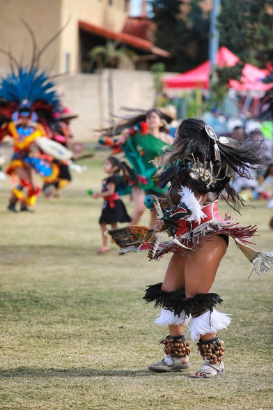 Aztec dancers wearing colorful ceremonial attire.  Large colorful headdress and intricate leather arm and chest pieces.  A young female dancer spins and her hair flairs out.  The skirt lifts out and the image captures her strong legs as she dances in front of others captured in the background through a soft bokeh.