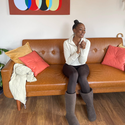 Brown leather couch with mustard yellow, and vinyl salmon pink square pillows. Faux leather brown couch. Woman with her head slightly turned to the right smiling. She is grabbing her right sleeve. She has on a cream/ white jacket, and dark brown pants, with dark brown boots.  