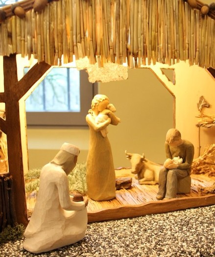 Die Weihnachtskrippe zeigt von links nach rechts einen knieenden König, dann Maria mit einem Lamm auf dem Arm, im Hintergrund ein Ochse und auf einem Baumstumpf sitzend Josef mit dem Jesuskind auf den Knien.