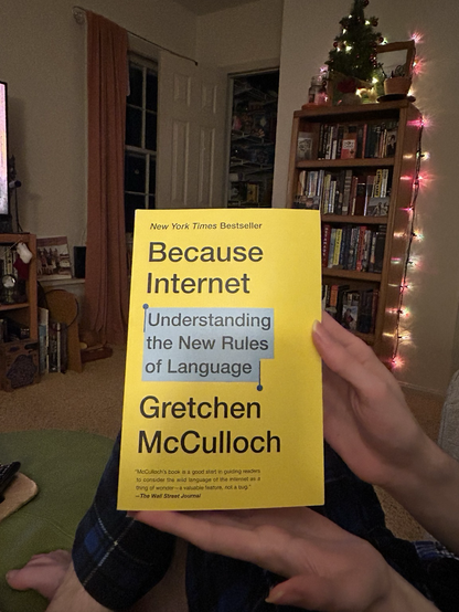 A hand holds the book Because Internet in front of a bookshelf decked out in Christmas lights.