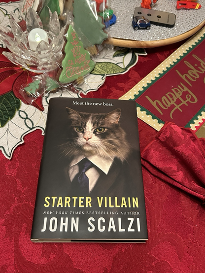 John Scalzi’s new book Starter Villain sits resplendently upon a cranberry red tablecloth, surrounded by cookies and a Holly table runner.