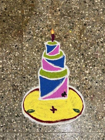 Colorful rangoli design of a birthday cake with a lit candle on a speckled floor.