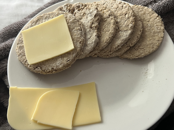 6 round shaped oatcakes on a white plate with a couple of slices of dairy free Gouda cheese. The plate is resting on a grey fuzzy blanket