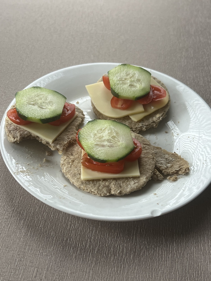 Three oatcakes on a white plate, on a muted gold table cloth. Each oatcake has non dairy Gouda cheese slice on them, some cut up cherry tomatoes, and a cucumber slice on the top