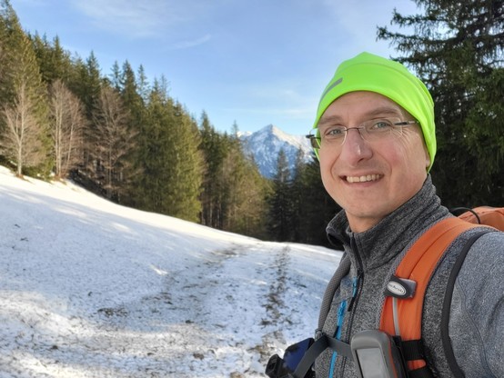This image captures a thrilling outdoor adventure scene. In the foreground, a man is taking a selfie against a wintry backdrop. He dons a pair of glasses and has a backpack strapped on, suggesting he is on a hiking expedition. The glimmer in his eyes and the smile on his face indicate his enthusiasm for the journey. Behind him, the grey sky and the snow-covered mountain suggest it's winter. A tree is also visible in the background, adding to the natural beauty of the setting. This picture also …