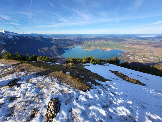This is a stunning image of a tranquil winter landscape. The primary focus is a snowy hill covered in snow, giving off a sense of calm and tranquility. The hill is surrounded by a vast expanse of nature, including trees and a serene lake that mirrors the clear, blue sky above. In the distance, a majestic mountain range creates a dramatic backdrop. The sky above is a vibrant shade of blue, dotted with fluffy white clouds. The scene is unspoiled and possesses an incredible sense of natural beauty…