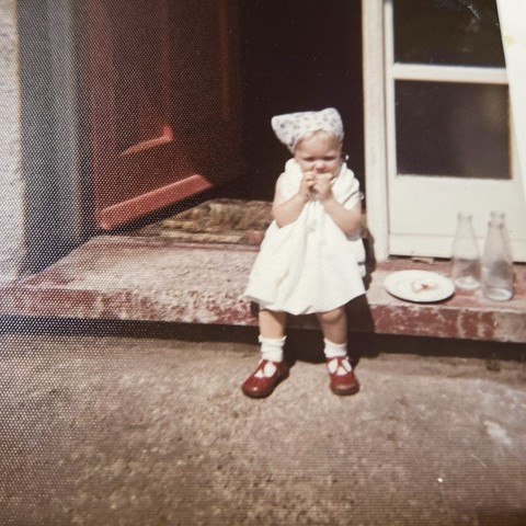 Square photo of me at about 18 months old. I’m sitting on the faded red step outside my maternal grandparents’ house in Nottingham. My passport was stamped at Heathrow in late April 1973 and this looks like the spring or summer since the red door to the left behind me is open and I’m wearing a white shapeless dress with white socks and red shoes. There is a white kerchief with an indiscernible pattern covering my short blonde hair. My hands are in front of my mouth and I’m eating what appears to be a cherry Bakewell tart. There is half a tart with the distinctive cherry and white icing sitting on a white plate on the step on the right side beside 3 empty glass milk bottles. Behind them are some windows with white framing. 