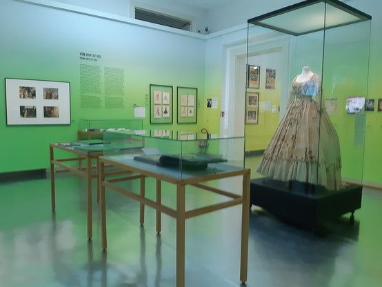 In hellem Grün gehaltener Ausstellungsraum. Im Hintergrund Informationen zur Entstehungsgeschichte der Sissi-Trilogie. Links im Vordergrund 2 Tische mit Ausstellungsstücken. Rechts hinter Glas ein hellbraunes Original-Kostüm aus einem der Sissi-Filme.