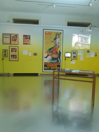 Ausstellungsraum. Im Hintergrund eine in sonnigem Gelb gehaltene Wand mit Filmplakaten, etwa "Spring Parade" mit Deanna Durbin und "Frühjahrsparade" mit Franziska Gaal. Rechts oben ein Monitor auf dem Ausschnitte aus "Die Deutschmeister" gezeigt  werden. Tisch mit Ausstellungsstücken im Vordergrund. 