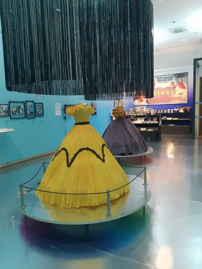 In hellem Blau gehaltener Ausstellungsraum. Im Vordergrund die Nachbildung eines gelben Kleides aus einem "Sissi"-Film. Dahinter ein nachgebildetes Kostüm mit dunkelgrauem Rock und dunkelgelbem Oberteil. Im Hintergrund ein Shop mit Büchern, DVDs, Kalendern u.a. zum Kaiserpaar Sissi und Franz Joseph. 